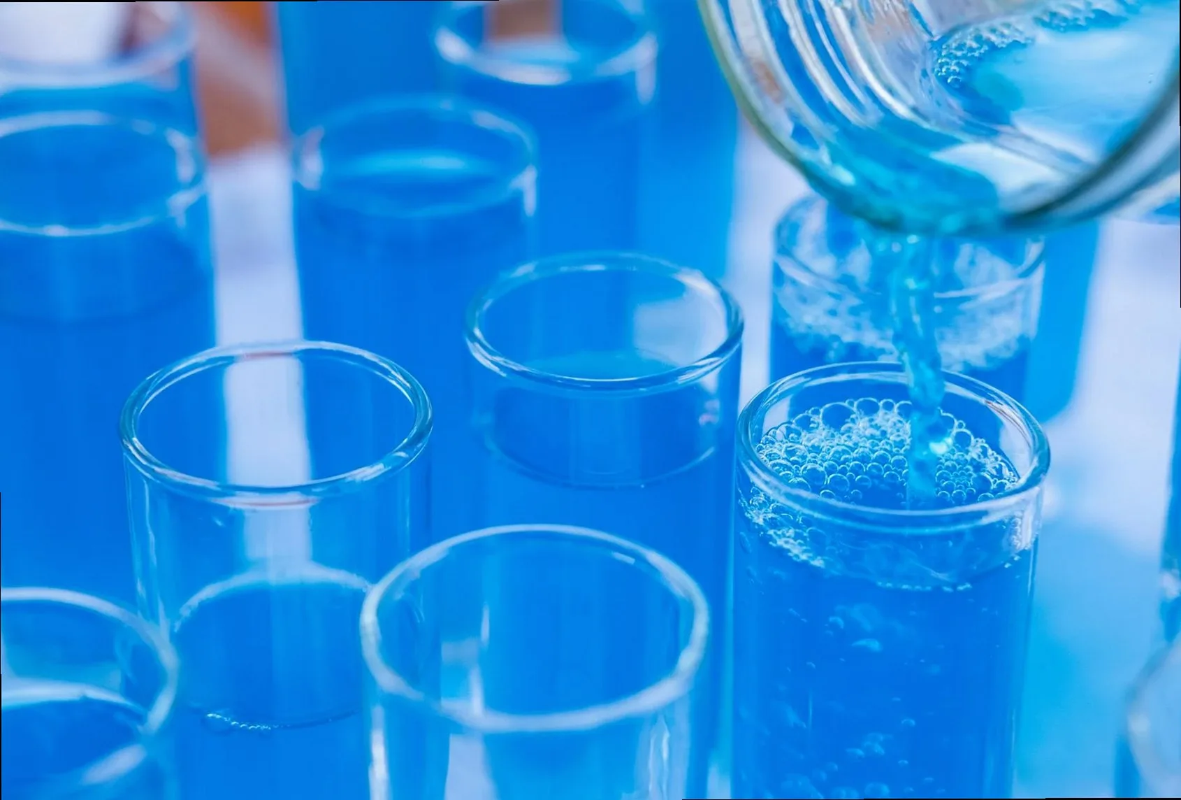 Close-up of blue liquid being poured into glass test tubes in a laboratory setting.